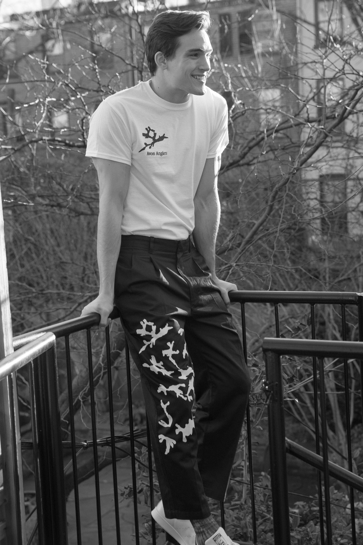 Smiling model wears Avon Anglers white tshirt with navy blot on the right chest with black logo embroidery, while perched on railing