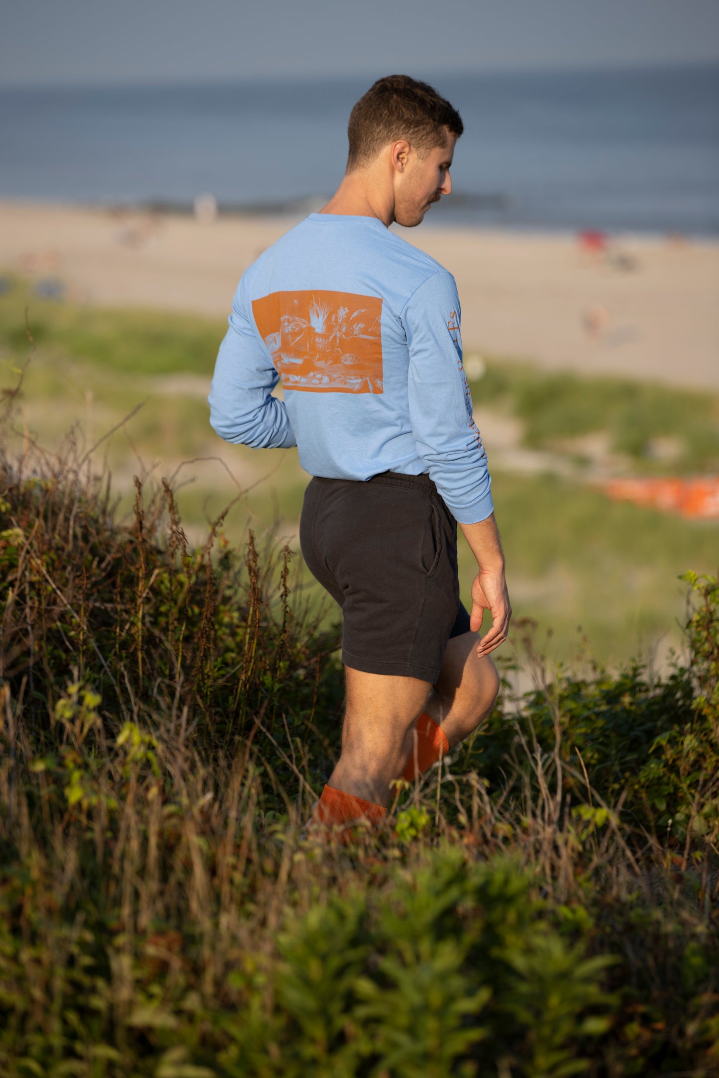 Model walking in sand dunes wearing Avon Anglers x Foppens long sleeve t-shirt in sky blue.  Back towards camera as he walks down the path