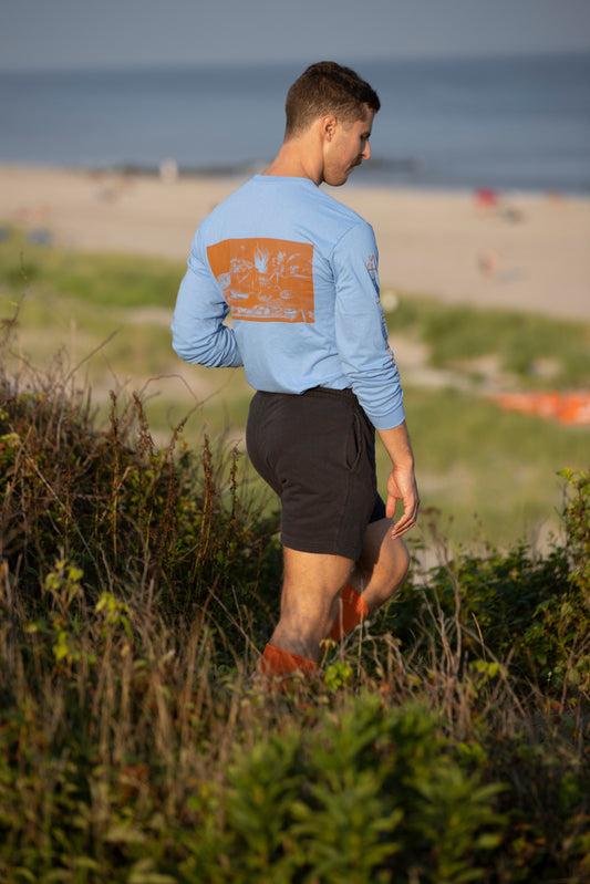 Model walking in sand dunes wearing Avon Anglers x Foppens long sleeve t-shirt in sky blue.  Back towards camera as he walks down the path