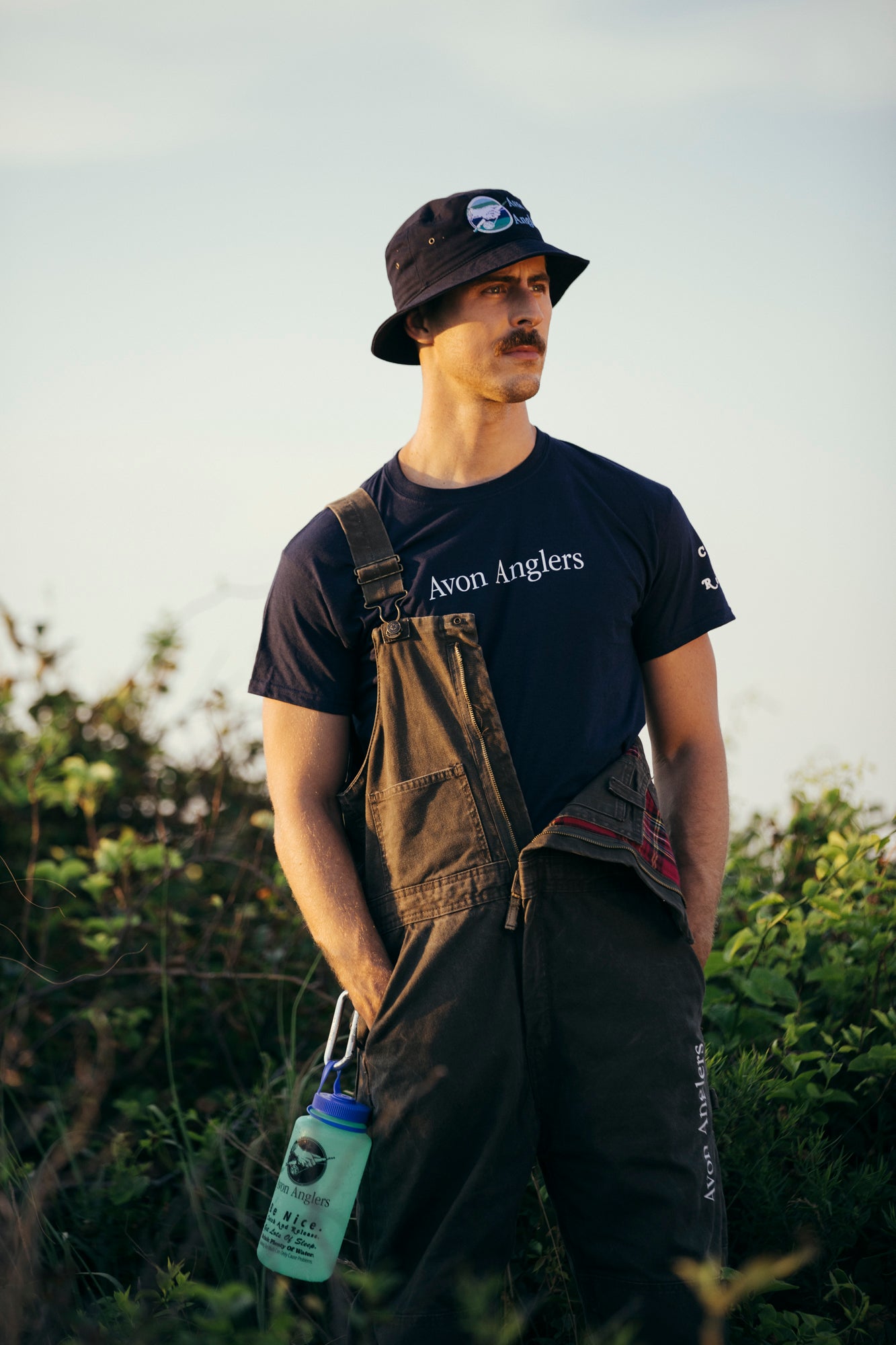 Model Nico Brown wearing Avon Anglers bucket hat, navy short sleeve t-shirt, re-wear bib front overalls with a Glow in the dark Nalgene bottle clipped to the coveralls