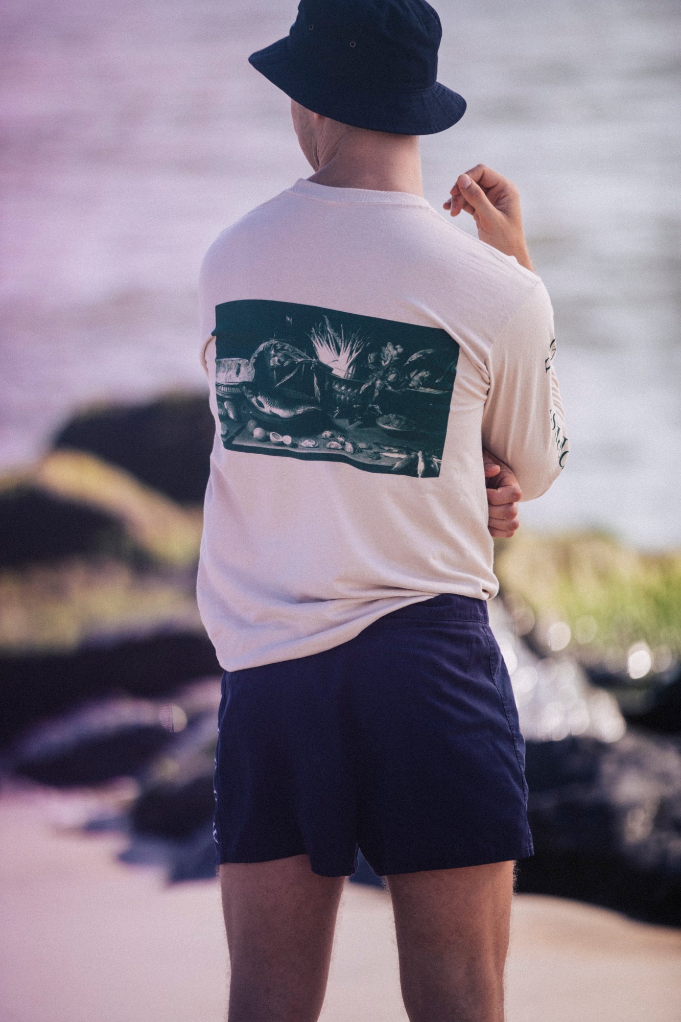 Model wearing Avon Anglers with his back to the camera looks out into the ocean wearing a sand colored long sleeve t-shirt with a Jacob Foppens Van Es print on the back of it.  He also wears a bucket hat and shorts