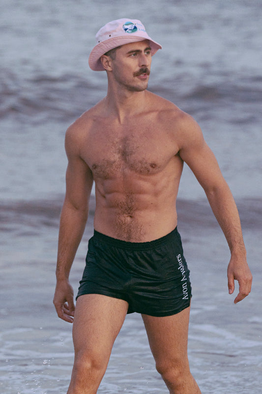 Model with a mustache and abs coming out of the ocean in a pink and rose colored bucket har.